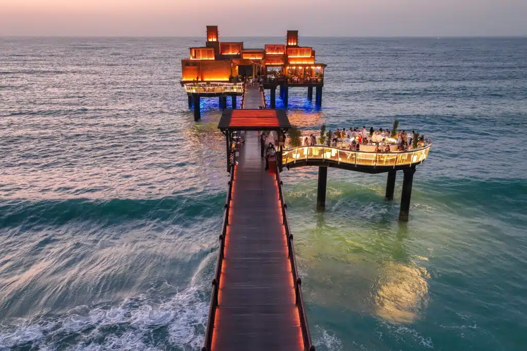 Dubai Best Restaurant Deals: A restaurant on stilts over the ocean at sunset with glowing lights, connected by a long pier.