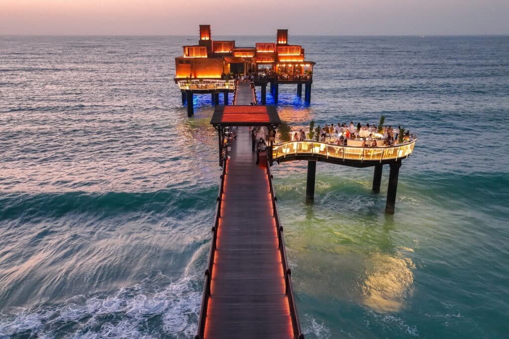A pier extends over the ocean at sunset, ending in a lit building and a circular platform with people.