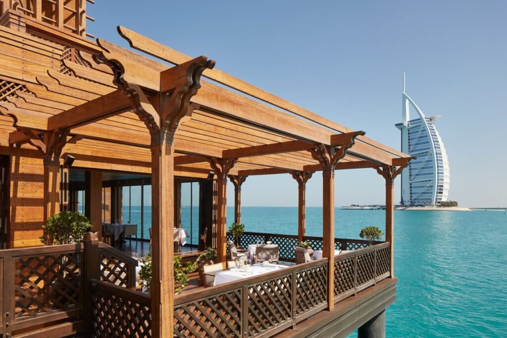 A restaurant terrace overlooking turquoise water with a distinctive sail-shaped building in the background.