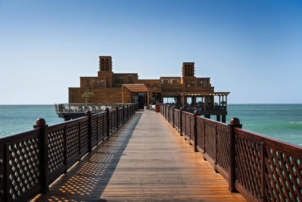 A wooden walkway leads to a building over the ocean, with clear blue skies and calm waters surrounding it.