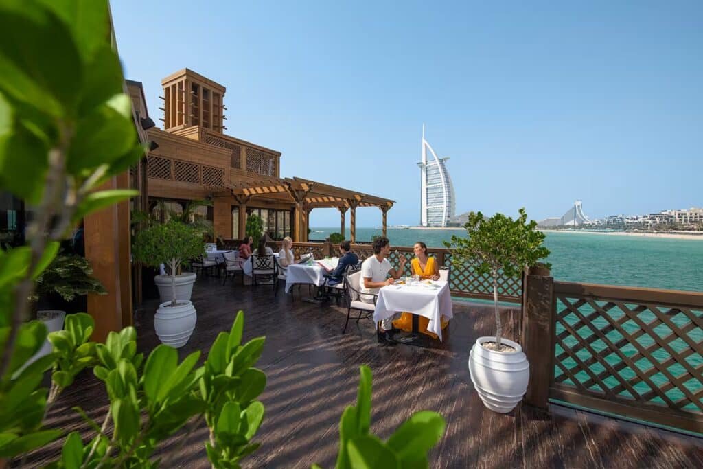 People dining on a wooden terrace overlooking the sea, with the Burj Al Arab in the background.
