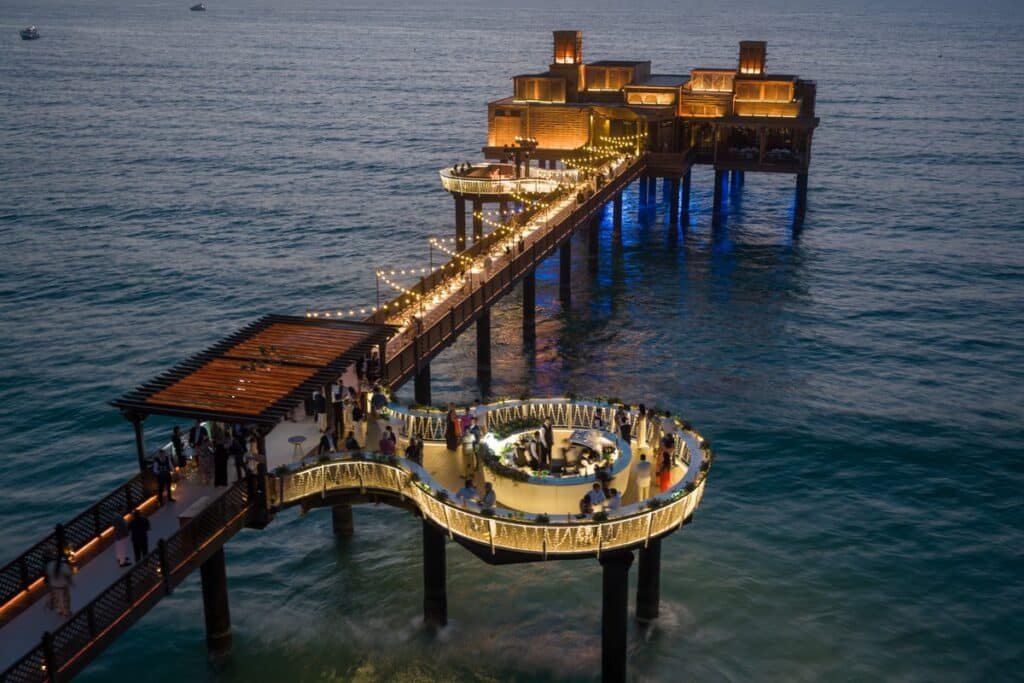 A lit pier extends into the ocean, featuring a curved Pierchic Dubai  dining area with string lights and a building at the end.