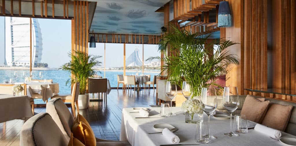Elegant Pierchic Dubai restaurant with panoramic ocean views, wooden decor, and tables set with white linens and glassware.