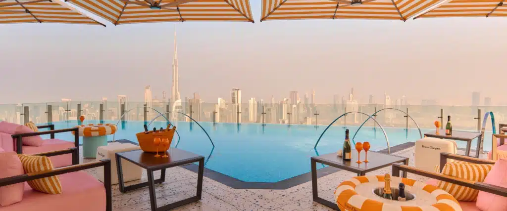 Infinity pool with orange and white loungers, overlooking a city skyline with a tall tower in the distance.