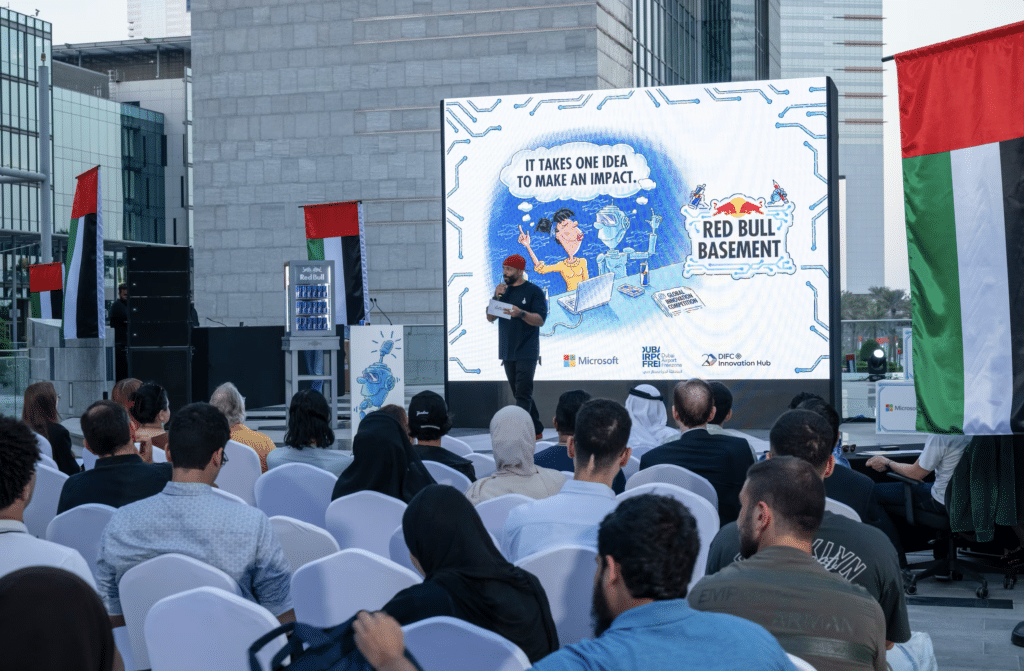 A speaker stands on stage addressing an audience, with a backdrop featuring a Red Bull Basement event poster.