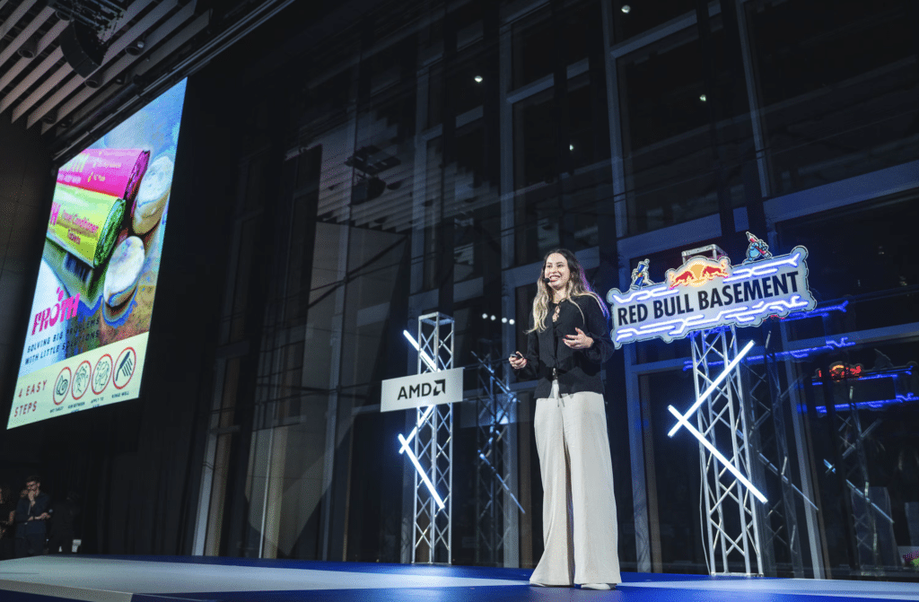 A woman stands on a stage giving a presentation with "Red Bull Basement" signage and an illuminated screen displaying a colorful slide.