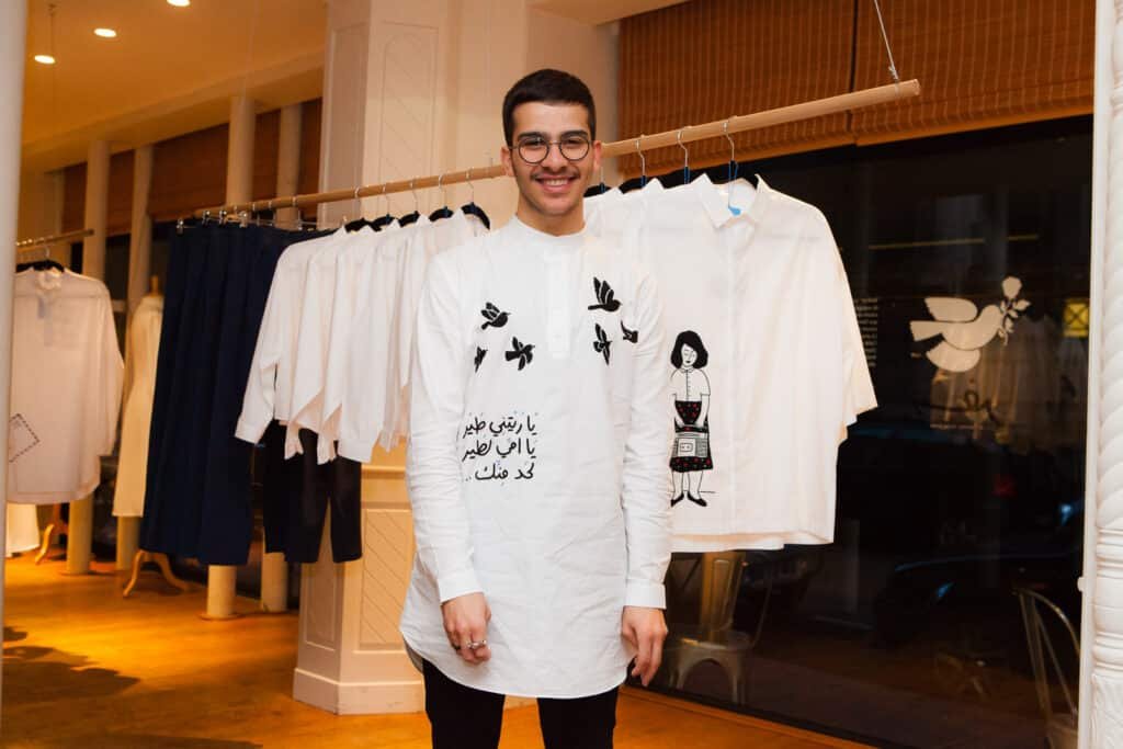 Support Lebanese Brands: Empowering Resilience, Craftsmanship, and Community from the UAE: A person wearing a white shirt with artistic designs stands in a clothing store with similar shirts on display in the background.