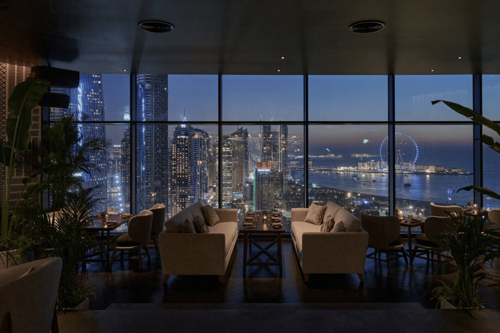 Elegant lounge with sofas overlooking a cityscape and coastline at dusk, featuring illuminated skyscrapers and a Ferris wheel.
