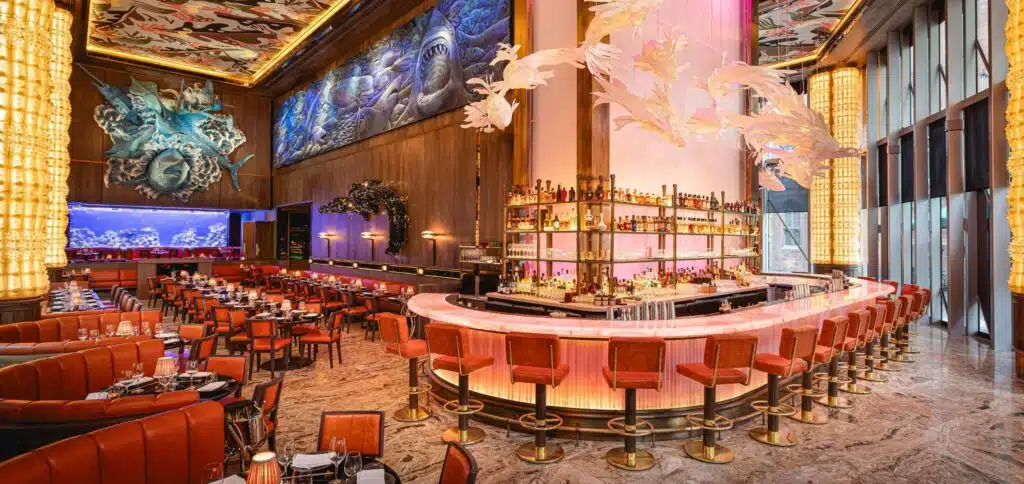 Elegant restaurant interior with a central illuminated bar, red leather seating, large wall murals, and artistic ceiling decor.