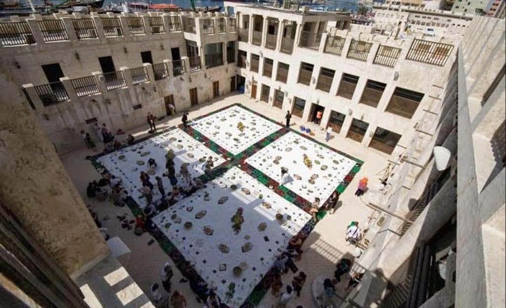 People gather in a large courtyard with patterned tiles and decorative elements, surrounded by multi-story stone buildings.