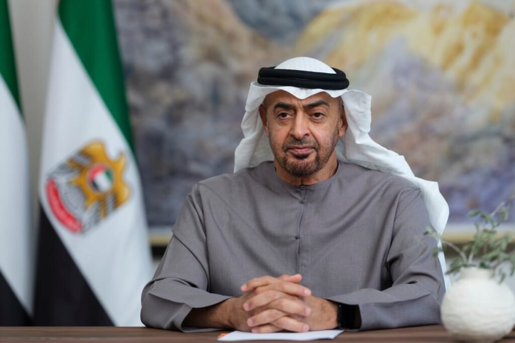 A man in traditional attire sits at a table with a UAE flag in the background.