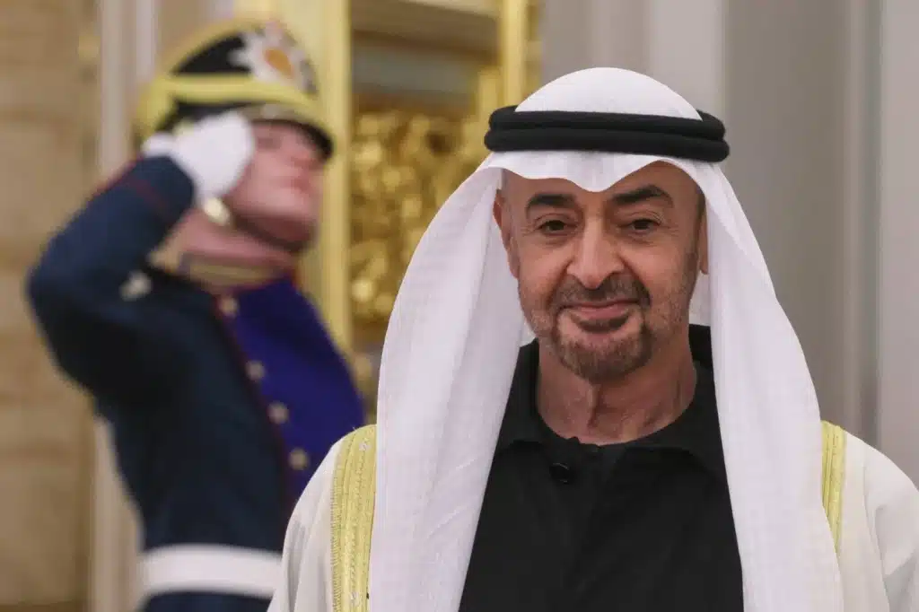 His Highness Sheikh Mohamed bin Zayed in traditional Middle Eastern attire stands in the foreground while a guard in formal uniform salutes in the background.