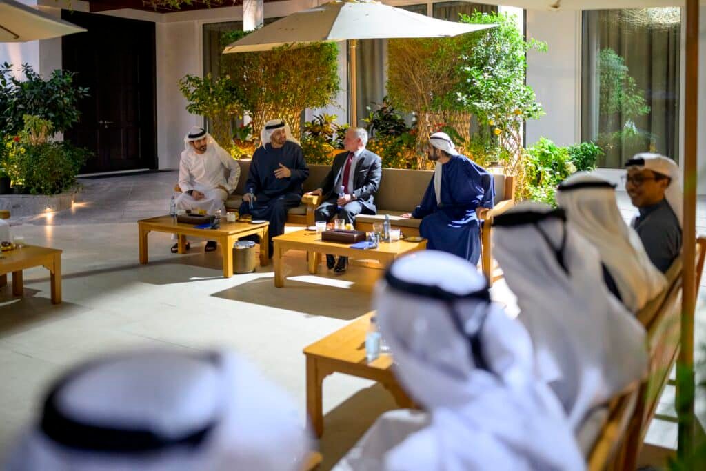 A group of people in traditional Arabic attire and suits sit around a patio table in a garden setting.