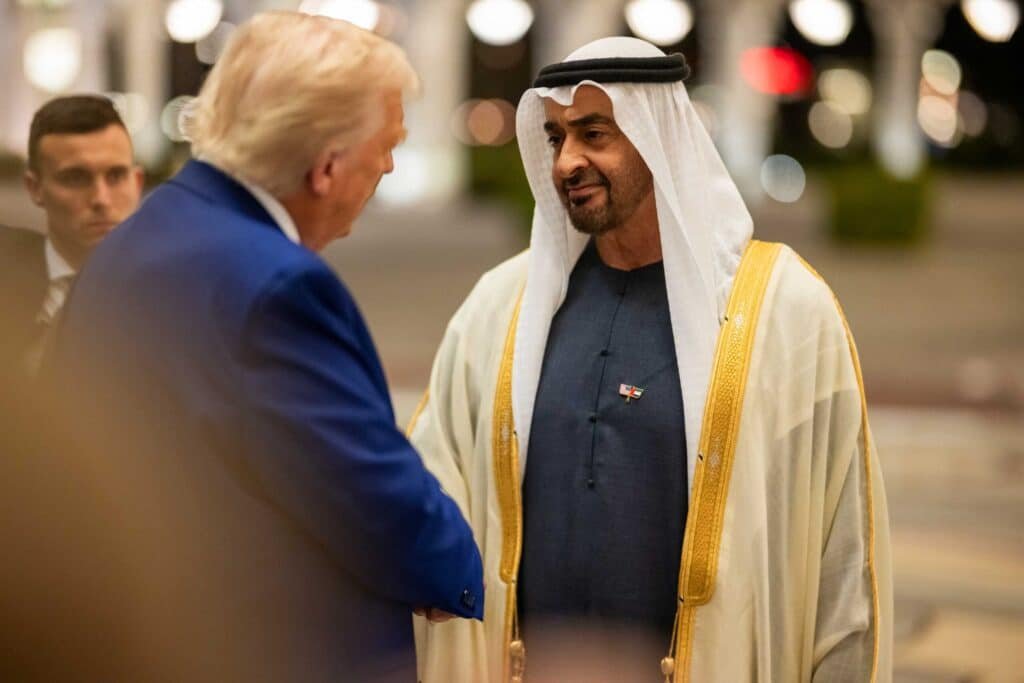 His Highness Sheikh Mohamed bin Zayed is shaking hands with Donald Trump; one wears a blue suit, the other a traditional white and gold robe, both appear to be in a formal setting.