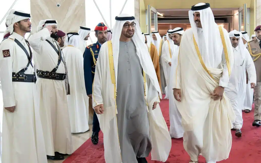 Two men in traditional white robes walk together on a red carpet, surrounded by a group of uniformed personnel.