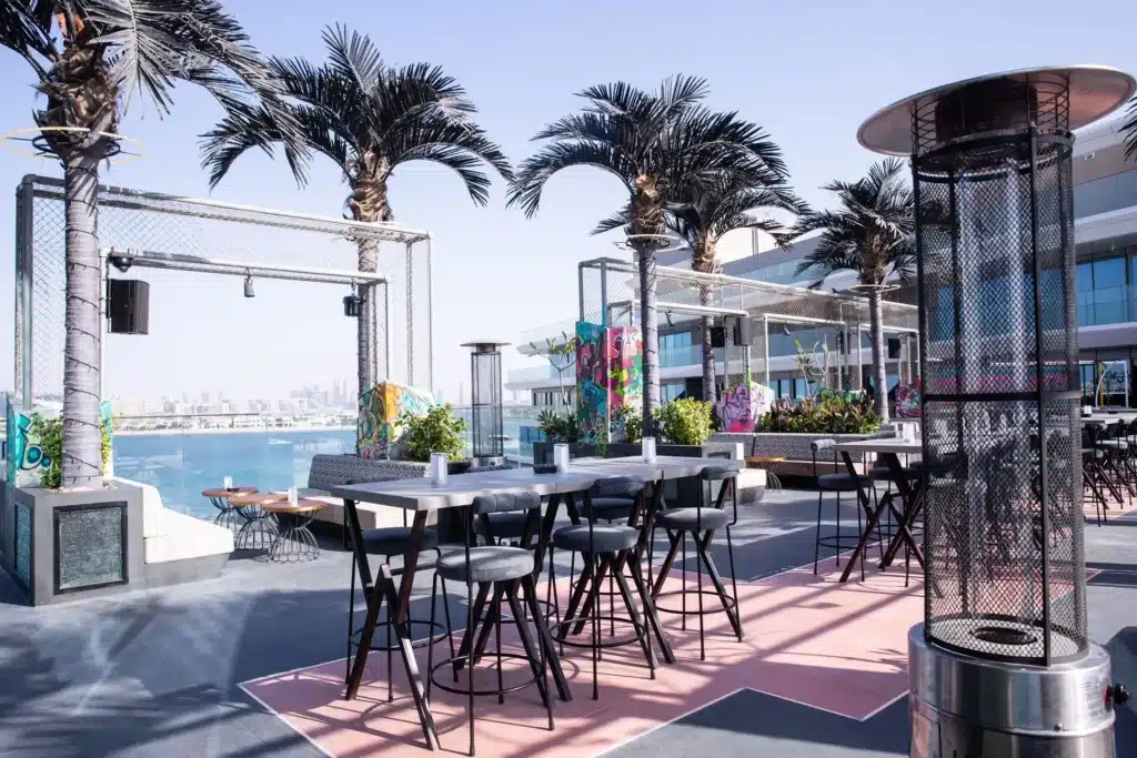 Rooftop terrace with modern furniture, palm trees, colorful murals, and a view of the city skyline in the background.