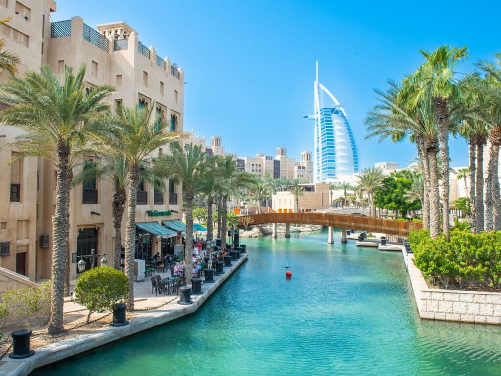 Scenic Souk Madinat canal with palm trees, traditional buildings, and the distinctive Burj Al Arab in the background under a clear blue sky.