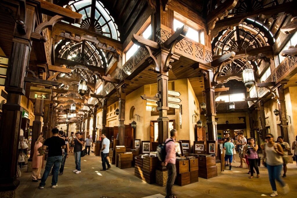 A bustling indoor Souk Madinat marketplace with wooden beams, ornate carvings, and various shops lining the walkway.