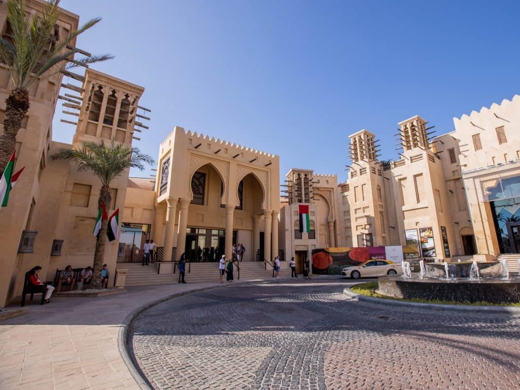 A Souk Madinat building with arched entrances and UAE flags, set against a clear blue sky.