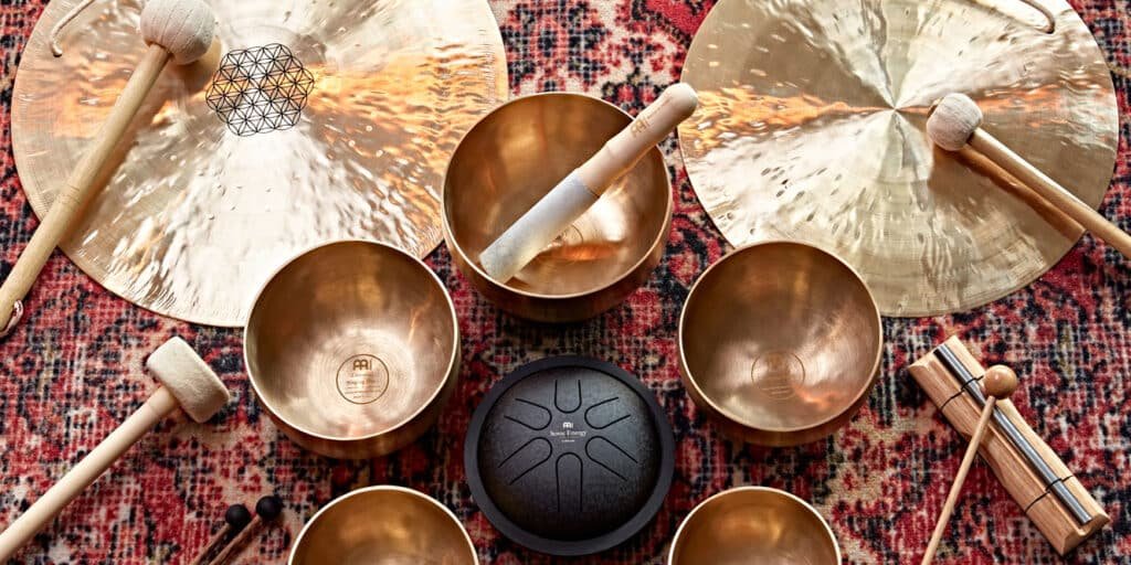 A variety of Sound Healing bronze singing bowls, two large cymbals, a black drum, and mallets arranged on a patterned textile.
