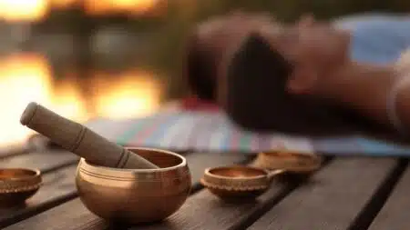A close-up of a singing bowl with a mallet on a wooden table, with two blurred people lying on a striped blanket in the background.