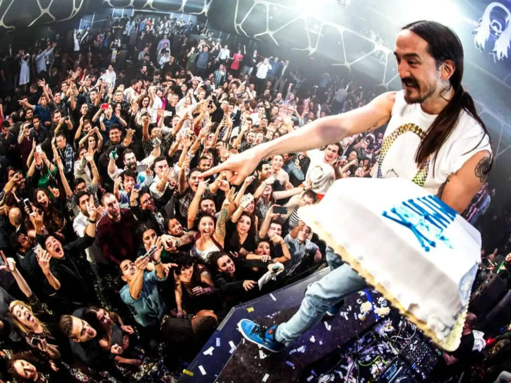 Steve Aoki Be Beach holds a large cake over a lively crowd in a nightclub, preparing to toss it.