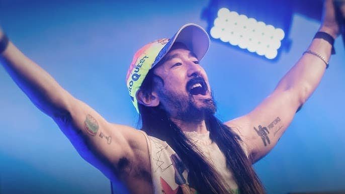 A man with long hair and tattoos on his arms raises his hands energetically under colorful stage lights.