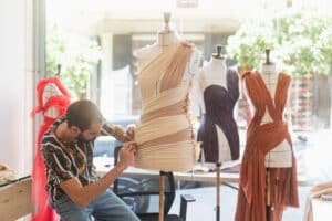 A fashion designer drapes fabric on a mannequin in a brightly lit studio with two other dressed mannequins nearby.
