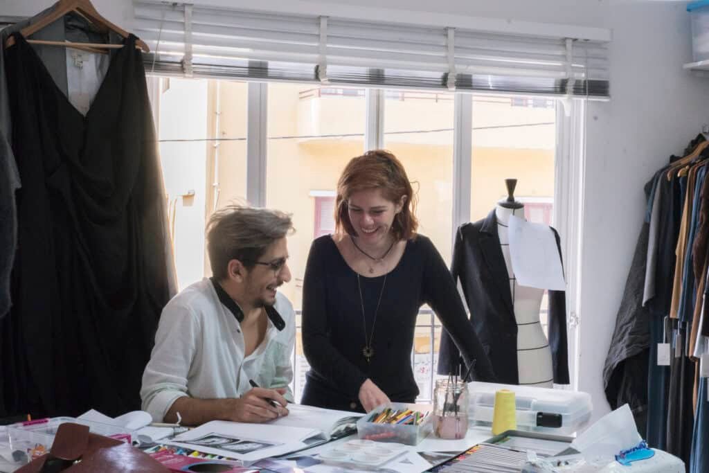 Support Lebanese Brands: Empowering Resilience, Craftsmanship, and Community from the UAE: Two people working at a cluttered table in a brightly lit fashion studio with a dress form in the background.