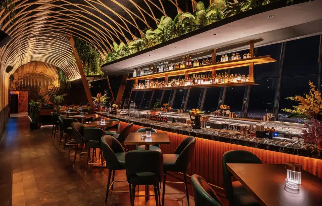 A stylish, dimly-lit bar interior with lush greenery, an arched wooden ceiling, and shelves of bottles behind the counter.