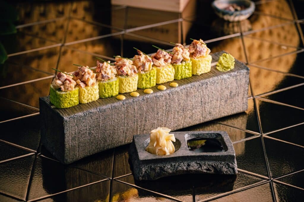 A row of gourmet sushi rolls with intricate toppings is presented on a stone platter, alongside wasabi and ginger on a smaller stone plate.