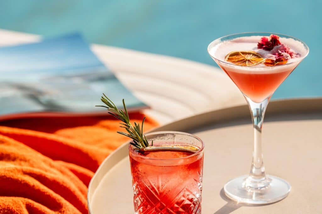Two cocktails on a tray by a pool, one in a martini glass with dried citrus, and another in a rocks glass with rosemary.
