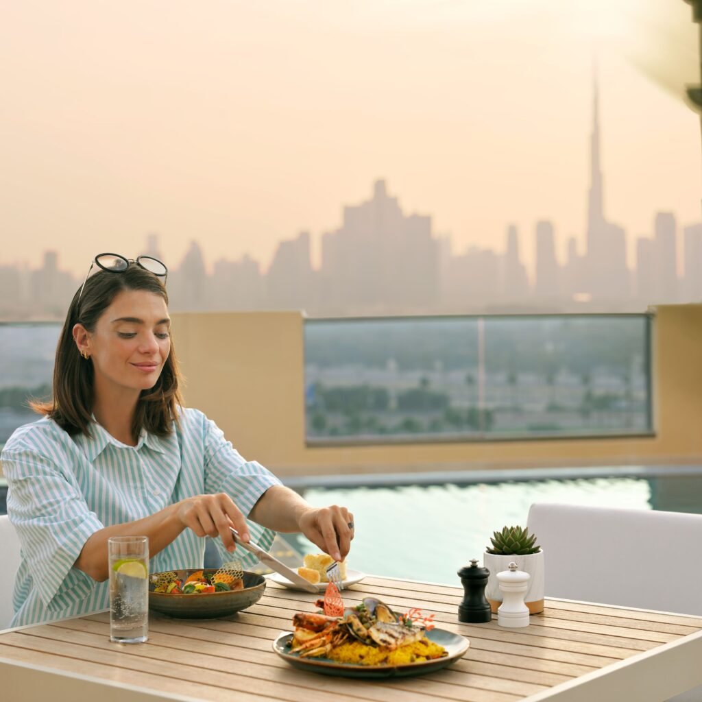 A woman enjoys a meal at an outdoor table with a city skyline, including a tall tower, visible in the background.