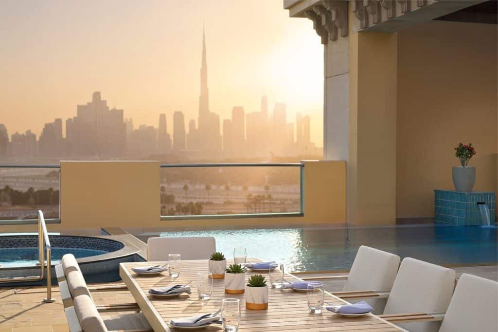 A rooftop dining area overlooks an infinity pool and city skyline at sunset, featuring a neatly set table with succulents and glassware.