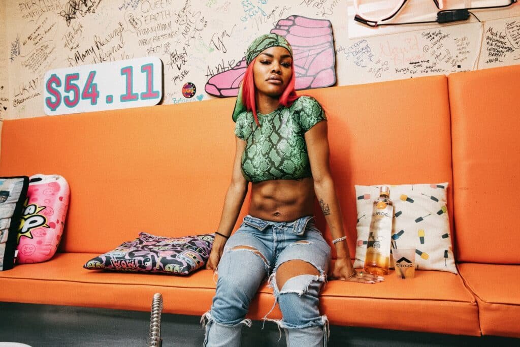 Teyana Taylor with red hair wearing a green crop top and ripped jeans sits on an orange couch surrounded by pillows and graffiti-covered walls.