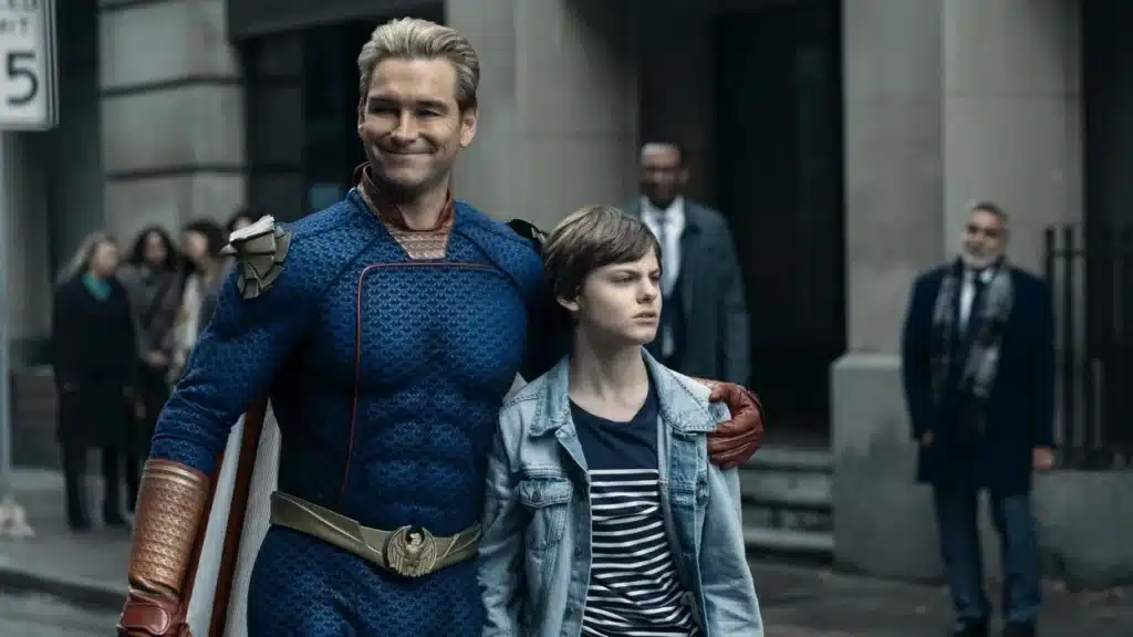 A man in a superhero costume with a cape smiles while walking on a city street with his arm around a young person.