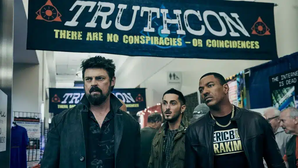 Three men stand under a banner reading  'Truthcon' in the tv show, The Boys Season 5