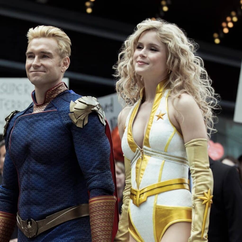 Two people dressed in superhero costumes walking with confident expressions. The person on the left is wearing a blue, armored suit with red and gold accents. The person on the right is wearing a white and gold outfit with a star emblem and long gloves.