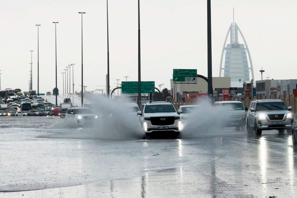 Cars drive through a flooded street, splashing water, with a distinctive triangular building in the background.