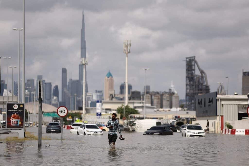 A man wades through a flooded street with partially submerged cars and buildings in the background.