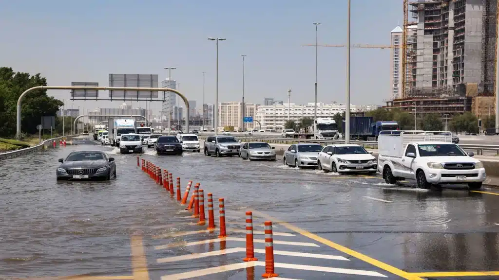 Cars driving through a flooded street with traffic cones and high-rise buildings in the background.