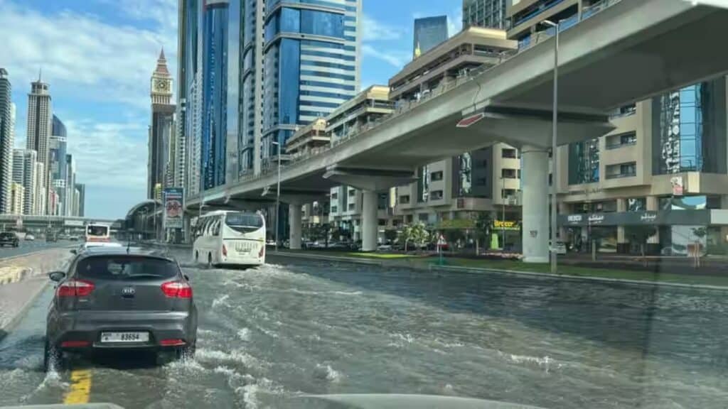 Cars driving through a flooded street in a city with tall buildings and an elevated roadway nearby.