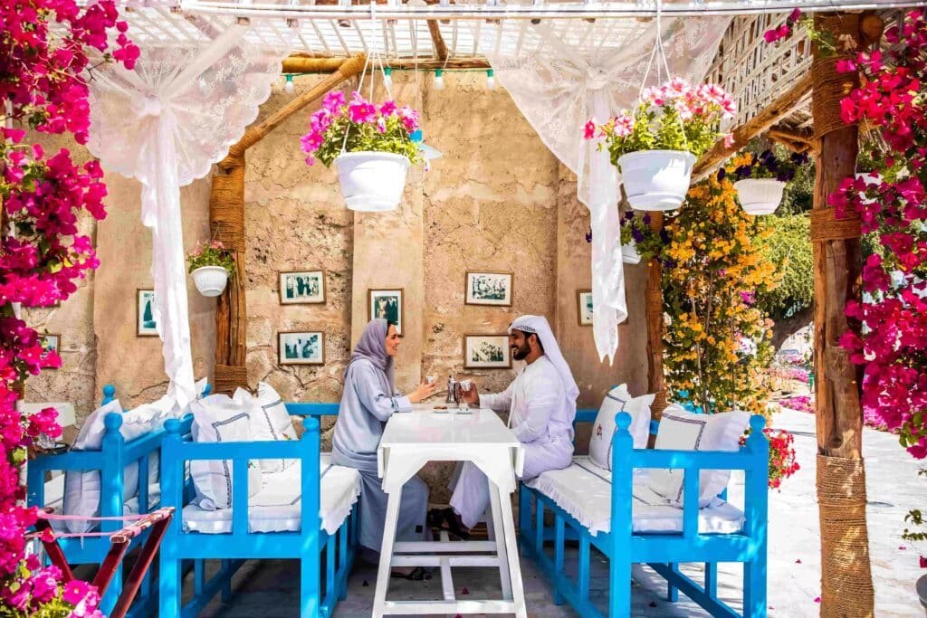 A couple in traditional attire sits at a blue outdoor café table surrounded by vibrant hanging flowers.