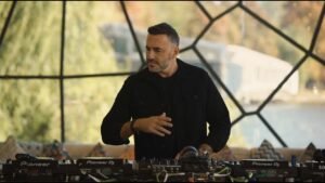 A DJ wearing a black jacket is performing at a DJ booth outdoors, with trees and a lake visible in the background.