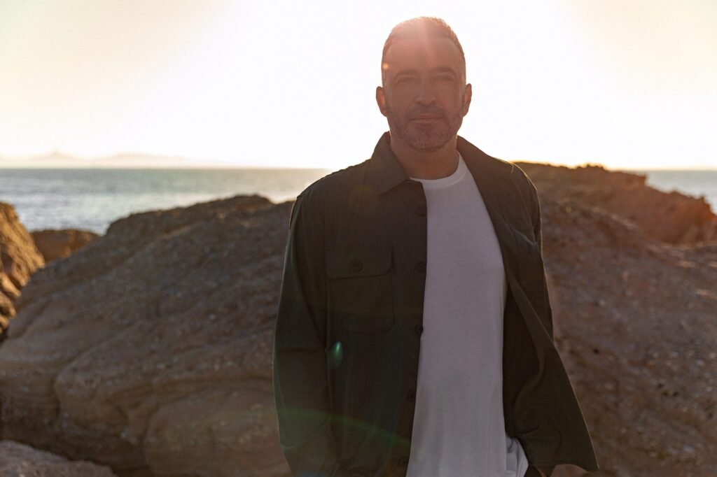 Valeron stands on rocky terrain by the sea with a soft sunlight glow in the background.
