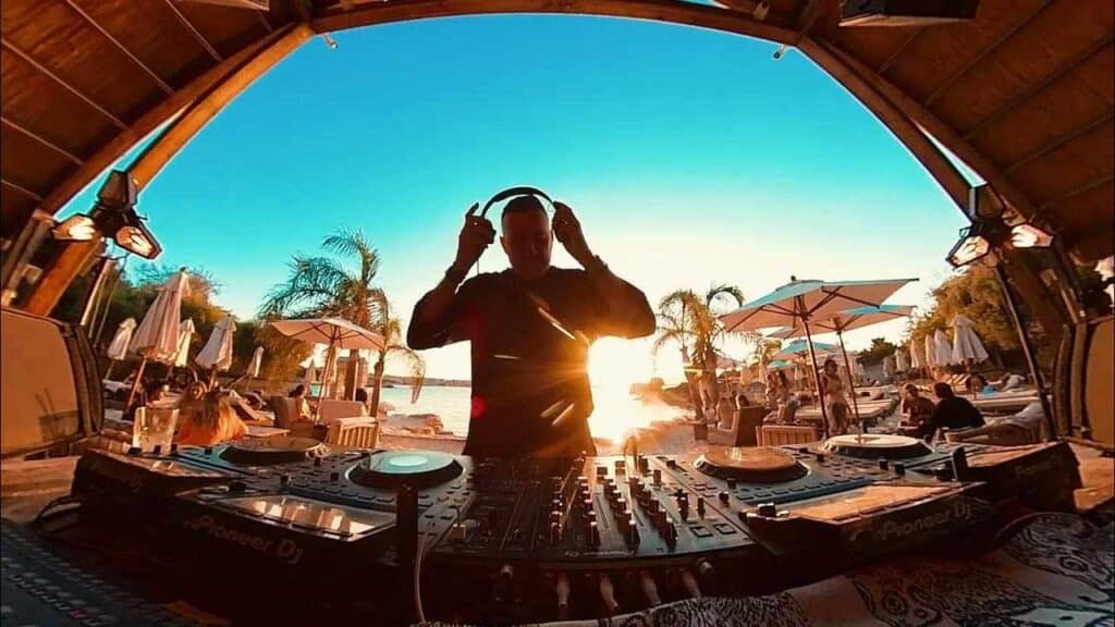 Valeron performing at a beachside venue during sunset, with turntables in front and palm trees visible in the background.