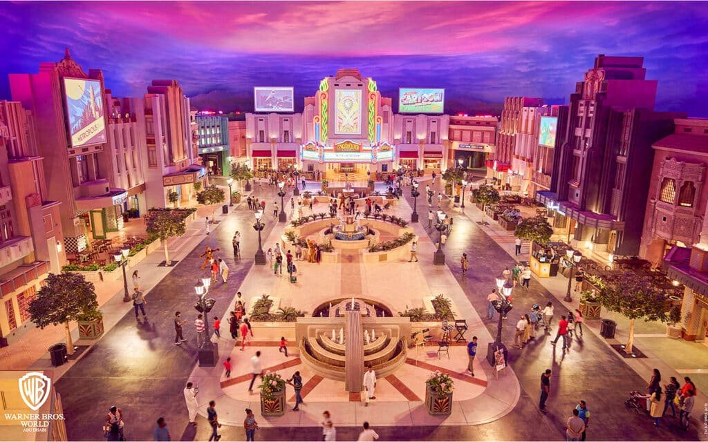 An aerial view of a themed amusement park plaza with art deco architecture, a central fountain, and visitors strolling around.