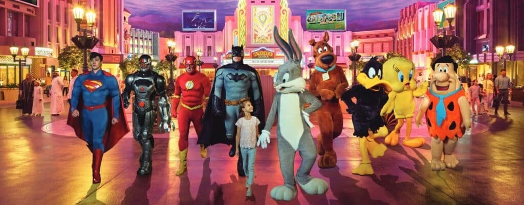 A child walks hand-in-hand with costumed characters of superheroes and cartoon figures in a brightly lit Warner Bros. World Abu Dhabi.