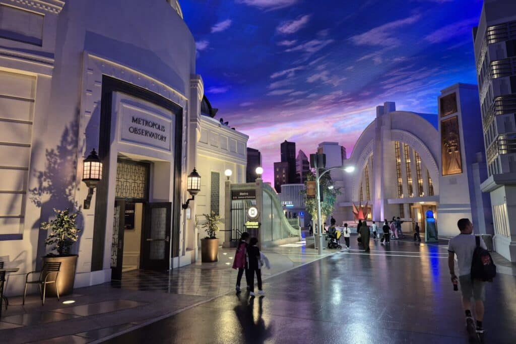 People walk along a street in a theme park, with Art Deco-style buildings and a sky painted on the ceiling.