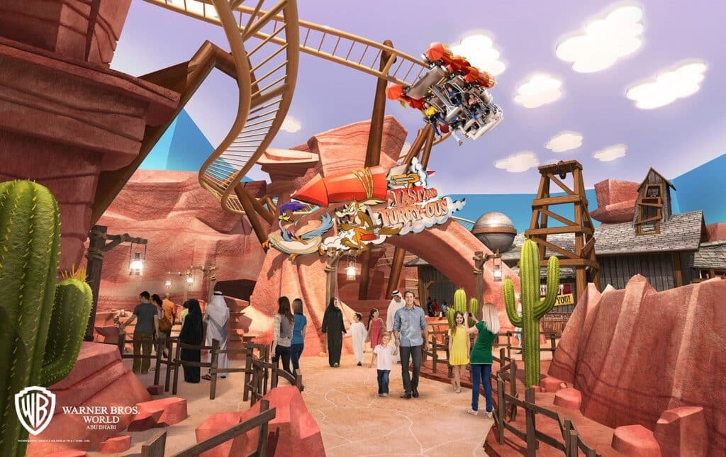 People walk under a roller coaster in Warner Bros. World Abu Dhabi area, surrounded by red rock formations and cacti.